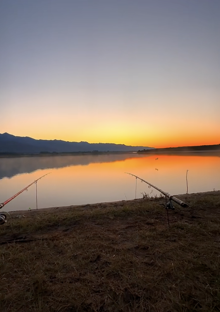Two fishing rods on a grassy bank beside calm water reflecting an orange sky, with hills along the horizon.