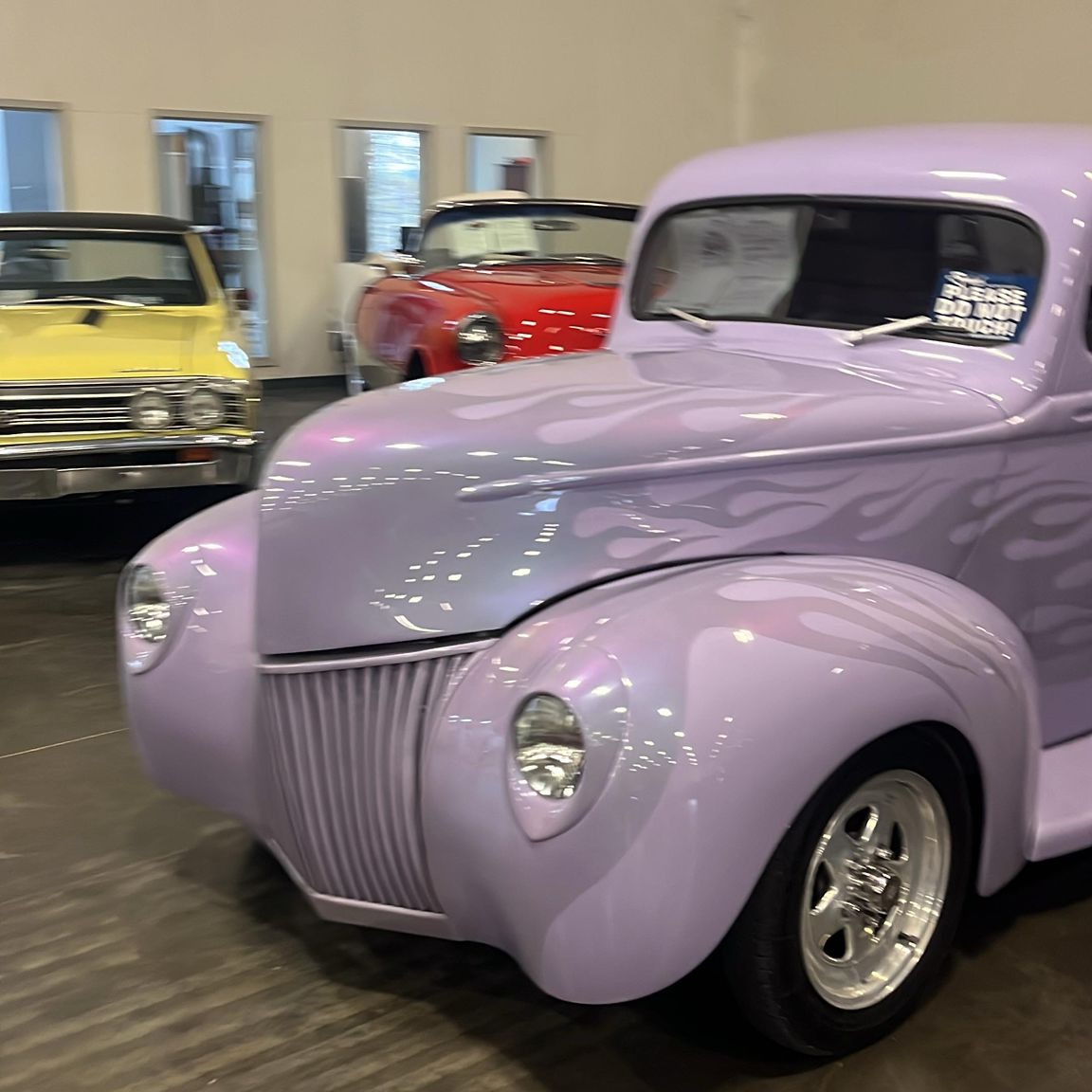 A lavender classic car with painted flames, displayed beside other vintage cars.