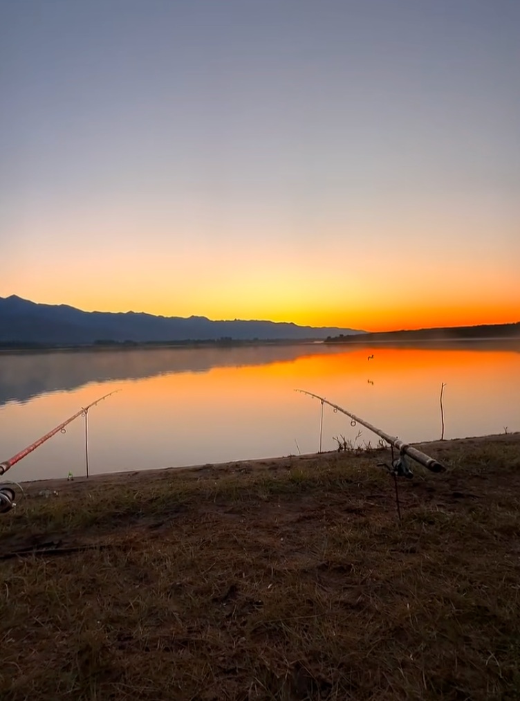 Two fishing rods beside still water reflecting an orange sky, with mountains on the horizon.